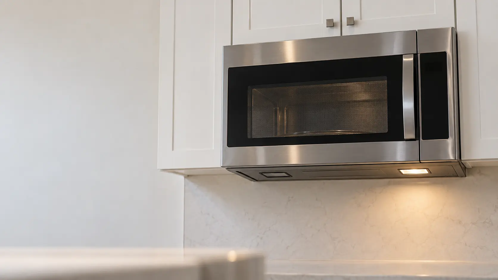 A stainless microwave hood combination under white wall cabinets seen from slightly below, one of the two small task lights on its underside dark and the other lit, its side-hinged door with a mesh window closed and the microwave cavity dim behind it.