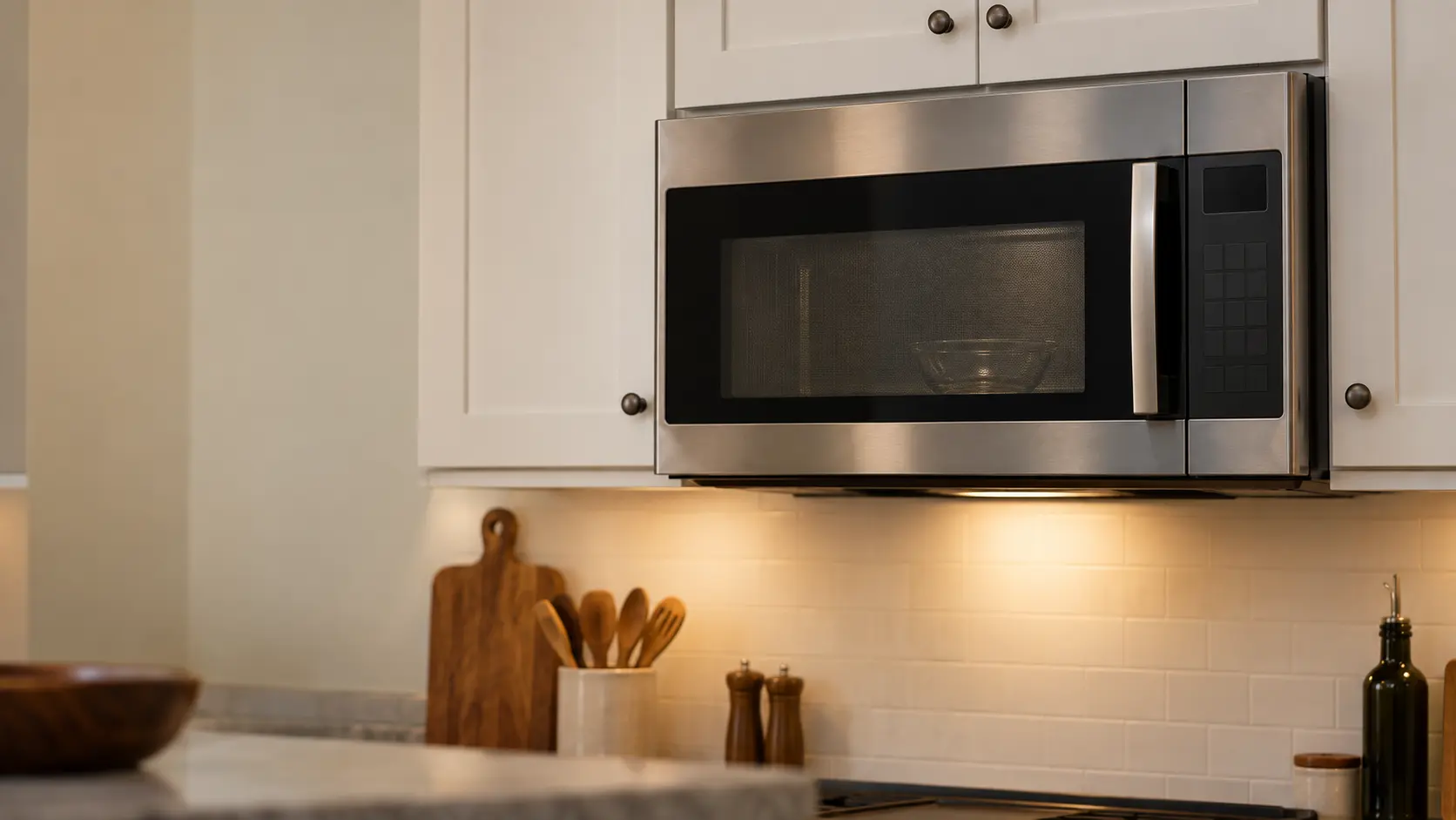 A stainless microwave hood combination mounted under white shaker wall cabinets in a US kitchen, the microwave's side-hinged door closed with its dark mesh window, a plain glass bowl visible faintly on the glass turntable inside the microwave, the lamp on its underside glowing softly onto a cooking surface that is cropped and out of focus at the bottom edge.