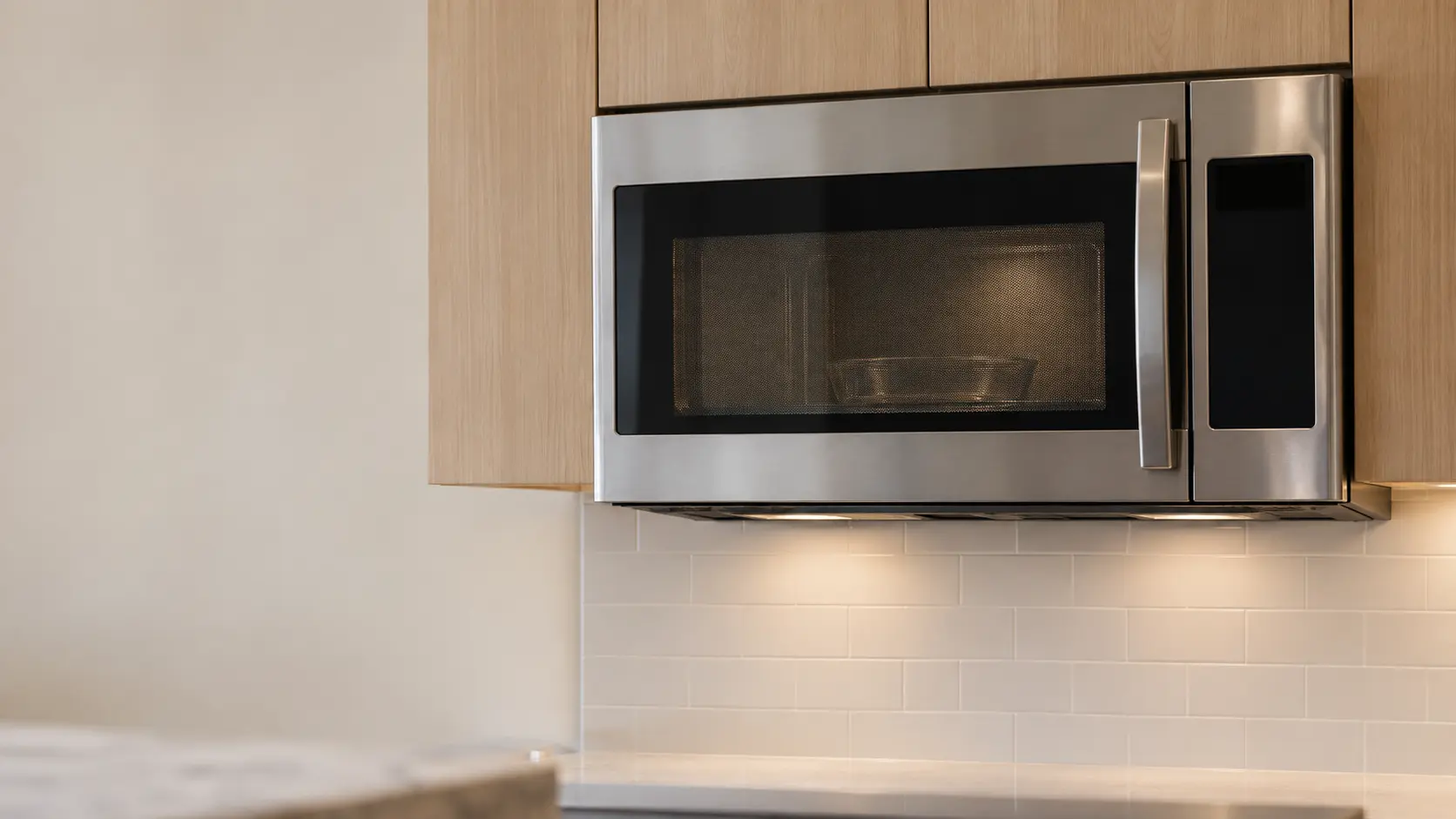 A stainless microwave hood combination freshly mounted under light wood wall cabinets in a home kitchen, its door closed with a plain glass dish on the glass turntable visible through the door window, the cooking surface below cropped and out of focus, a clean tiled backsplash with a gap below the microwave, the display blank.